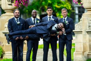 Groomsmen in navy suits playfully carry groom horizontally in garden setting.