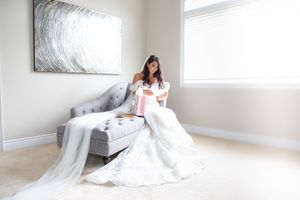 Bride in white gown opens gift while seated on gray chaise lounge.