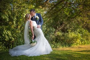 Bride and groom share a romantic kiss outdoors surrounded by lush green trees.