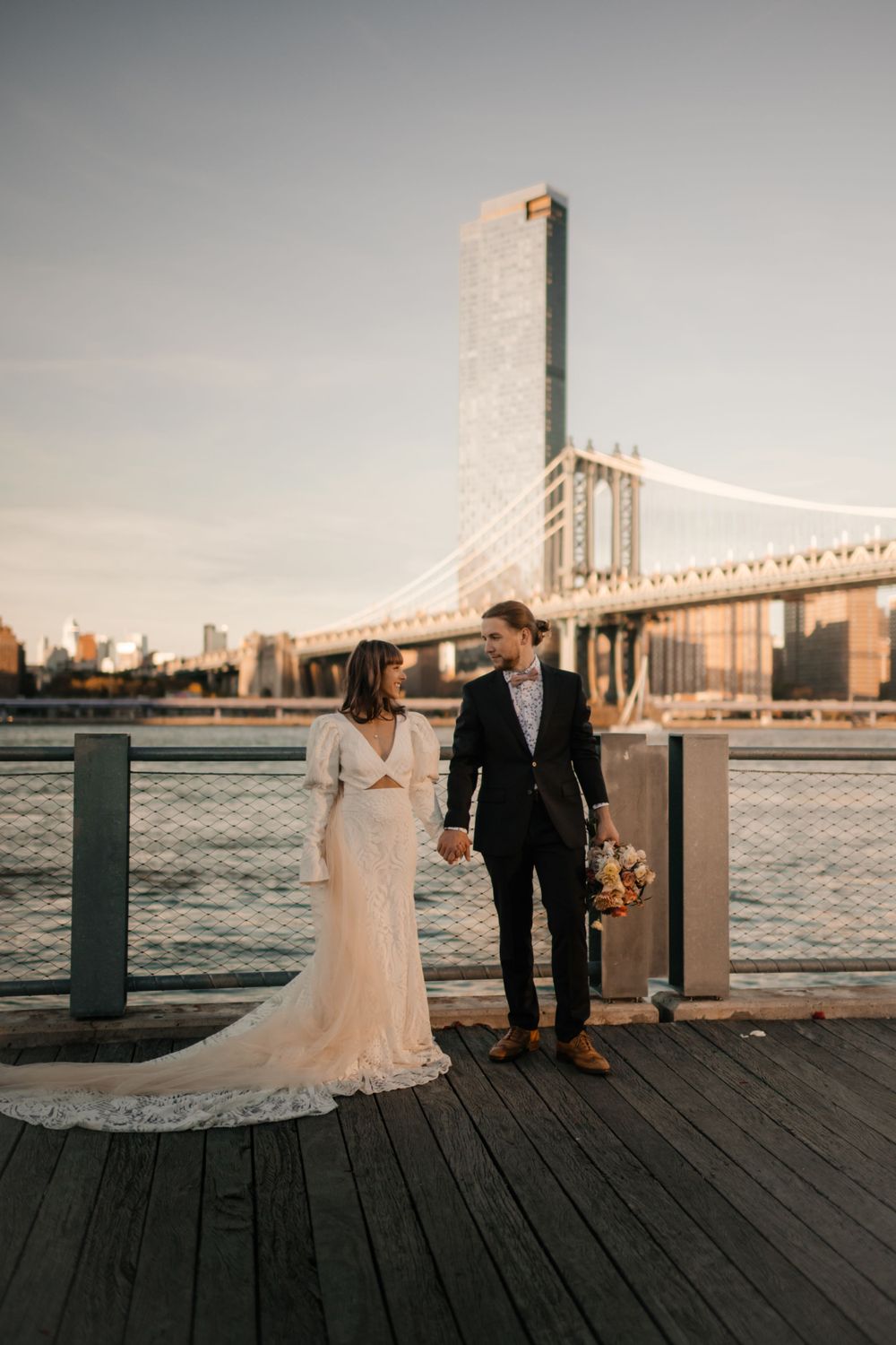 Newlyweds holding hands on boardwalk with Manhattan Bridge and NYC skyline in background.