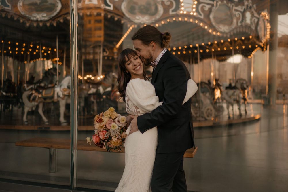 Newlyweds embrace in front of illuminated carousel during evening wedding photoshoot.
