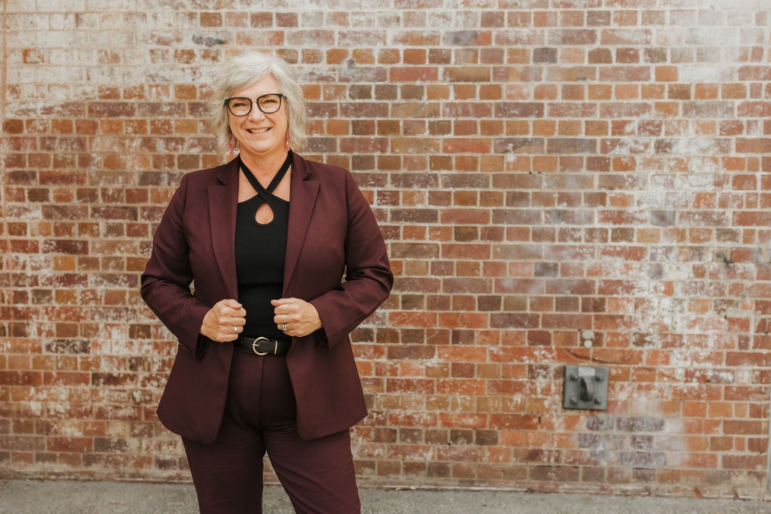 Professional wearing burgundy suit and glasses standing against brick wall.