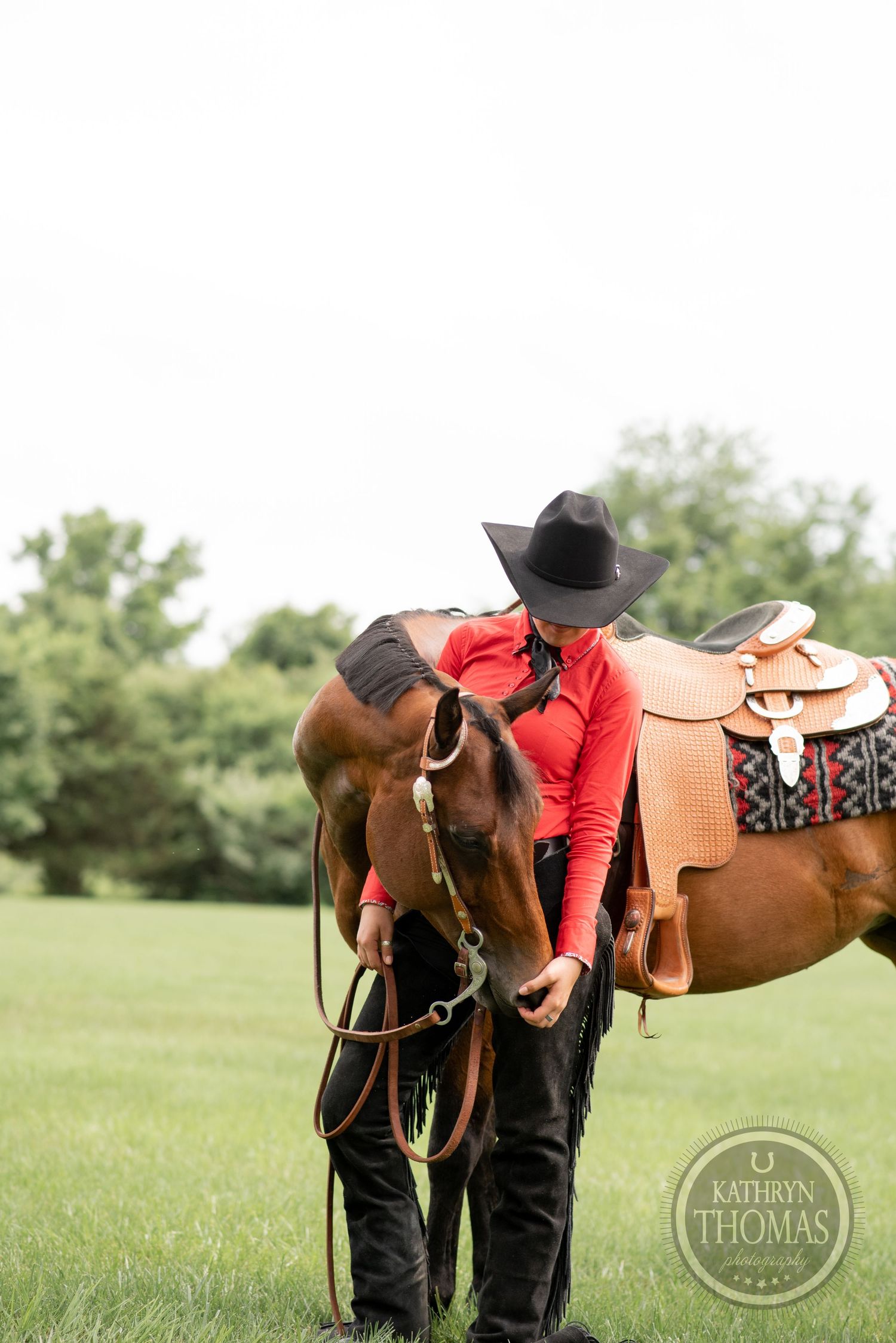 Person in red shirt and cowboy attire stands with western-saddled horse in grassy field.