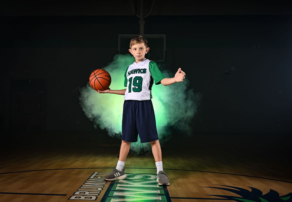 Youth basketball player poses with ball amid dramatic green smoke effects on court.