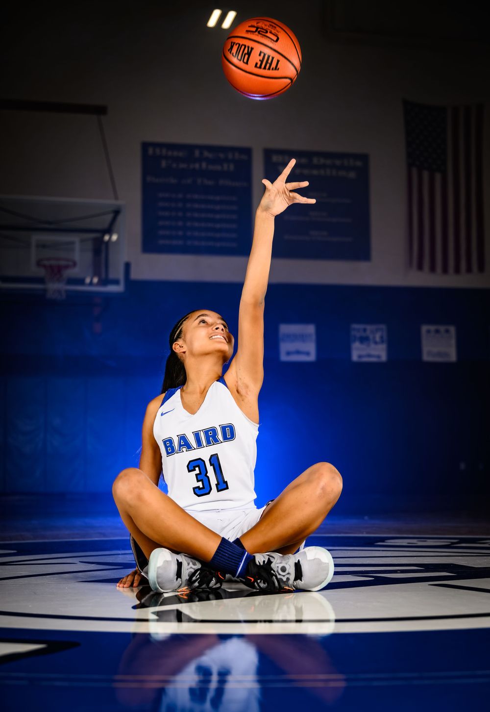 Basketball player in white jersey sits on court tossing ball upward with dramatic lighting.