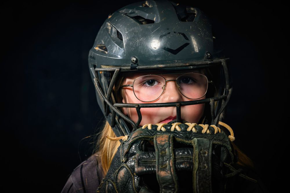 Close up portrait of softball player wearing protective gear and helmet against dark background.