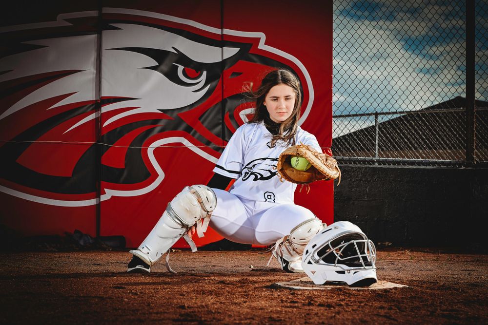Softball player poses in front of team mascot mural with equipment on dirt field.