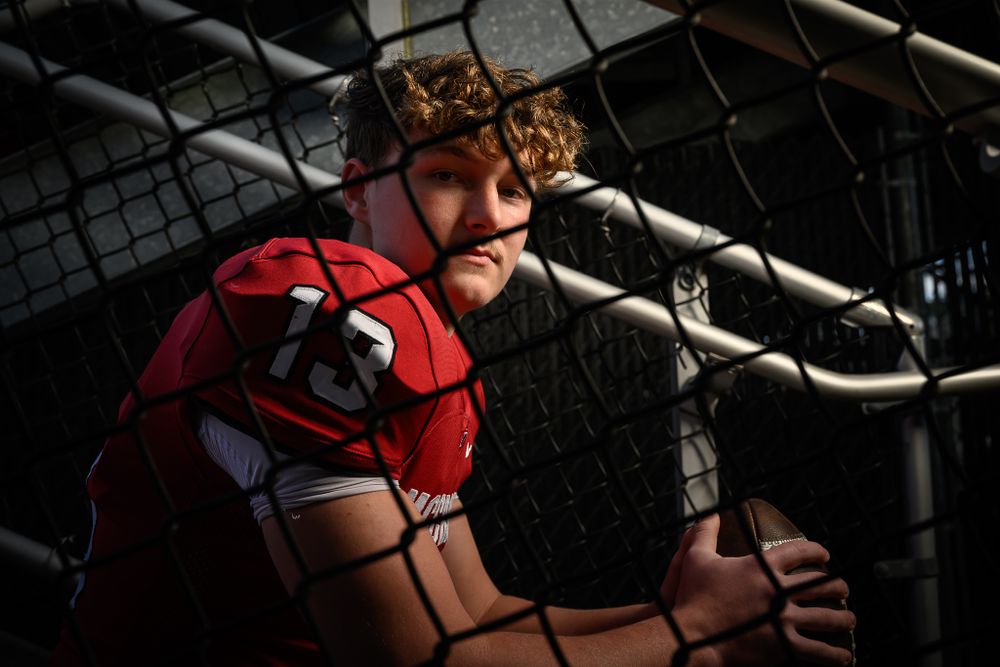 Baseball catcher in red gear crouches in dramatic lighting behind fence.