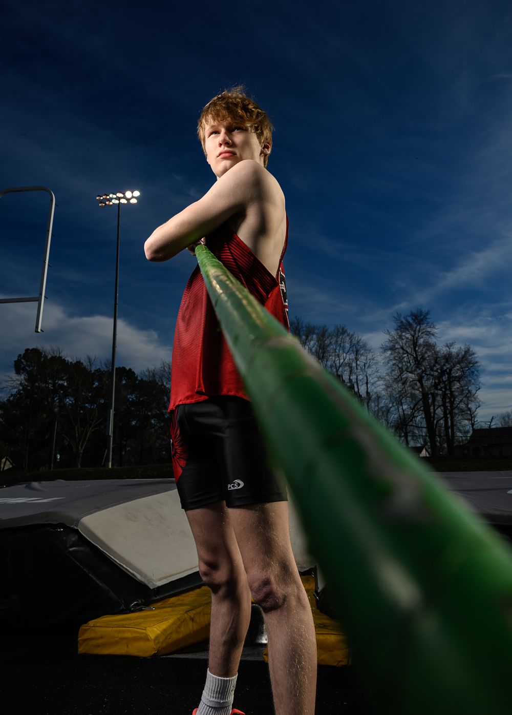 Athletic figure in red uniform holds a green pole against deep blue sky with stadium lights in background.