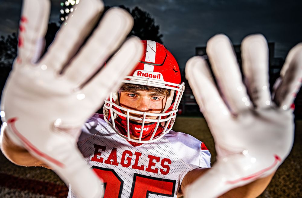 Football player in red Eagles uniform with helmet holds hands up to block the camera in dramatic close-up.