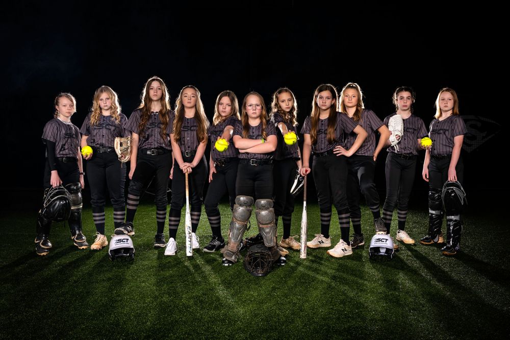Group of softball players pose together in black uniforms on field at night.