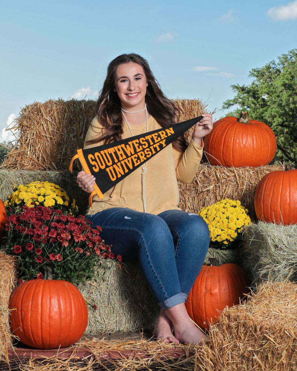 A brunette female student sitting on hay bails, pumpkins, and flowers holding a Southwestern University banner.