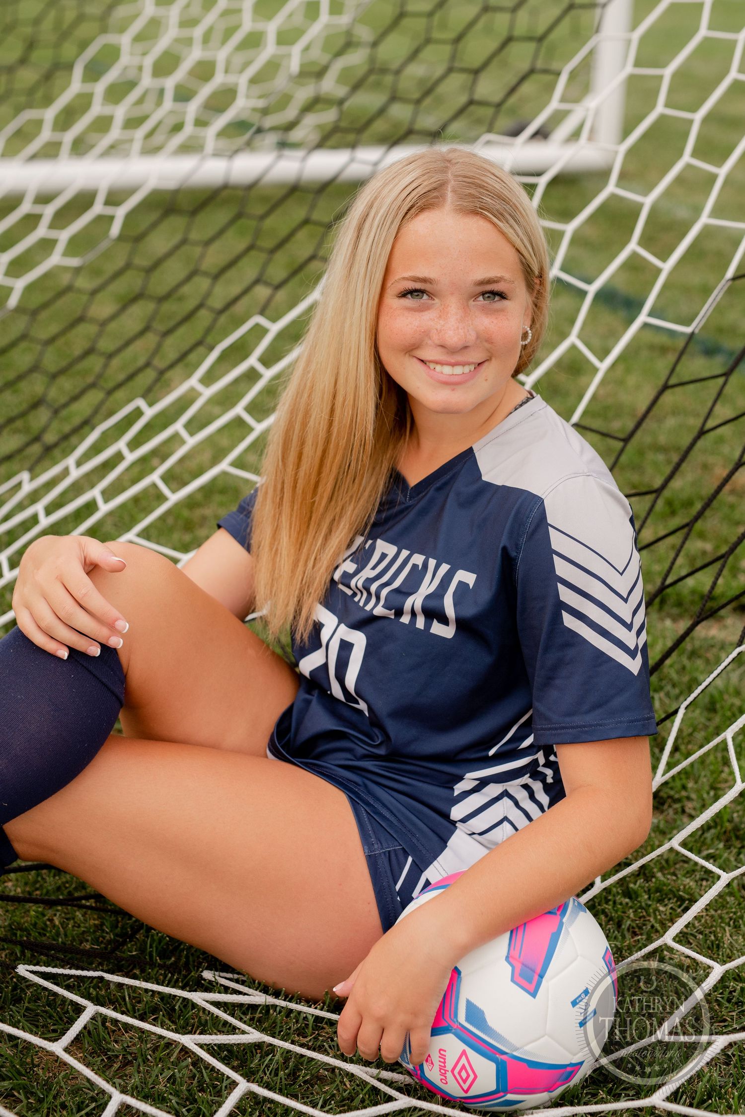Soccer athlete in navy Maverick jersey sitting with ball in goal net.