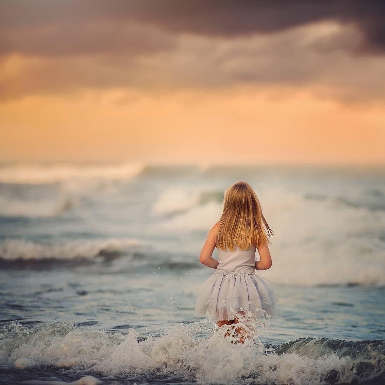 Blonde girl in white dress standing in ocean waves at dramatic sunset shoreline.
