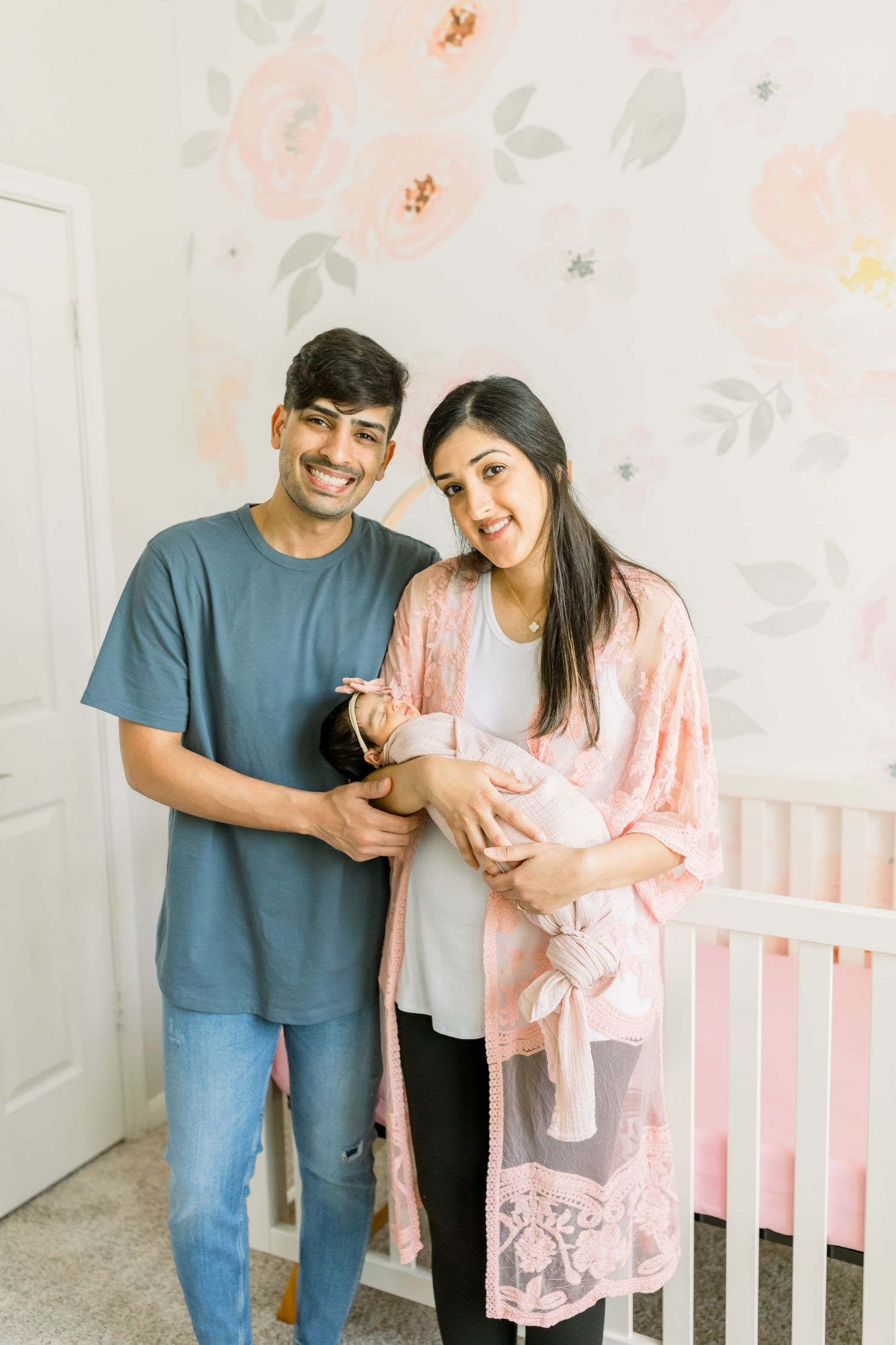Parents pose with their newborn baby in front of floral wallpaper and pink crib.