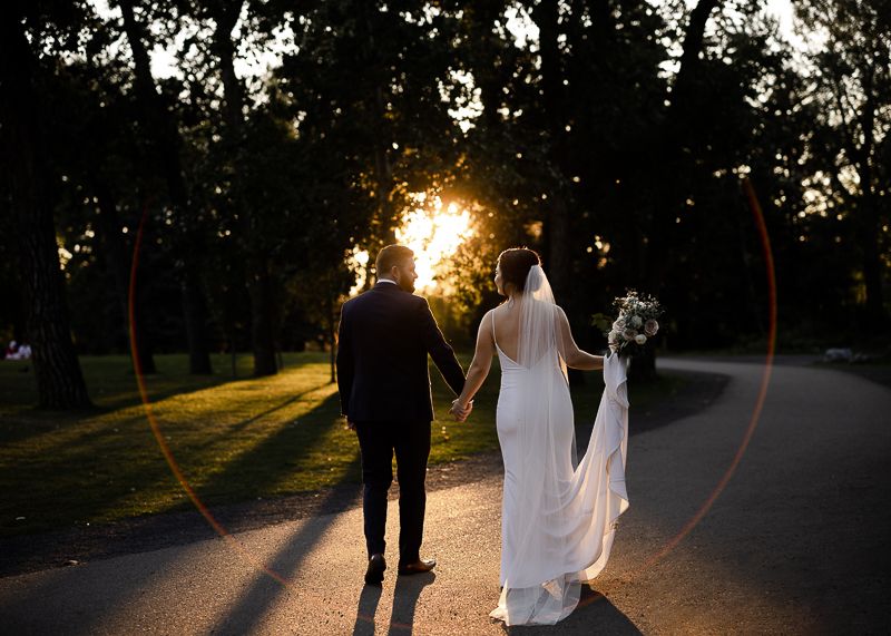 Sunset creates long shadows as newlyweds walk through park with lens flare.