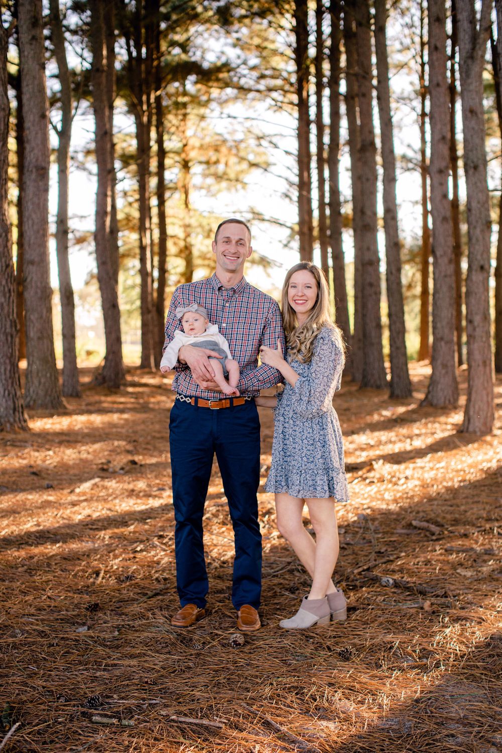 Outdoor family portrait in a sun-dappled pine forest with warm autumn lighting.