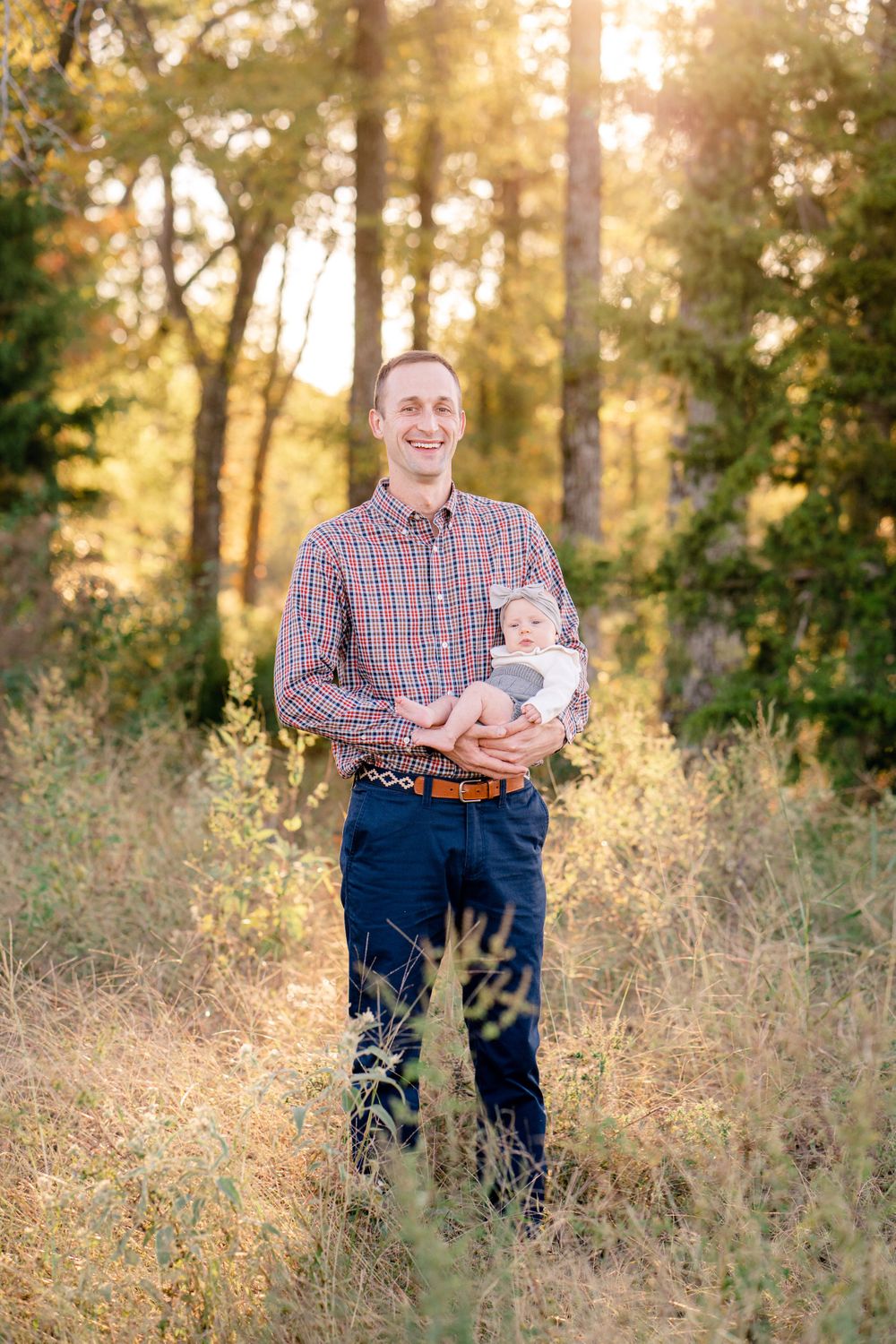 A golden sunset illuminates a wooded clearing as an adult holds a baby surrounded by tall grass and trees.