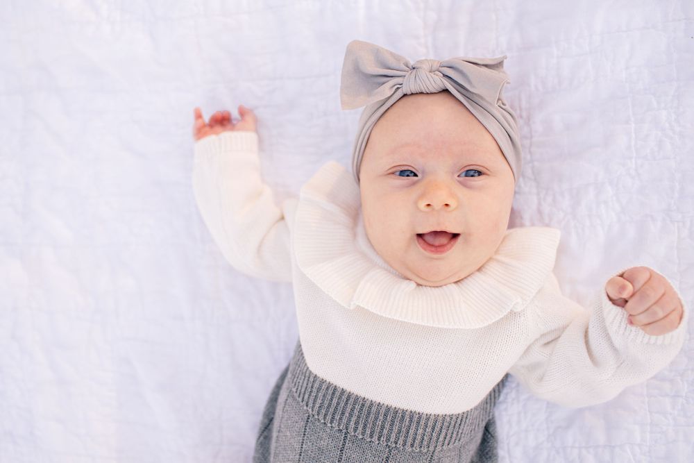 Baby wearing grey bow headband and white sweater smiles while lying on white blanket.