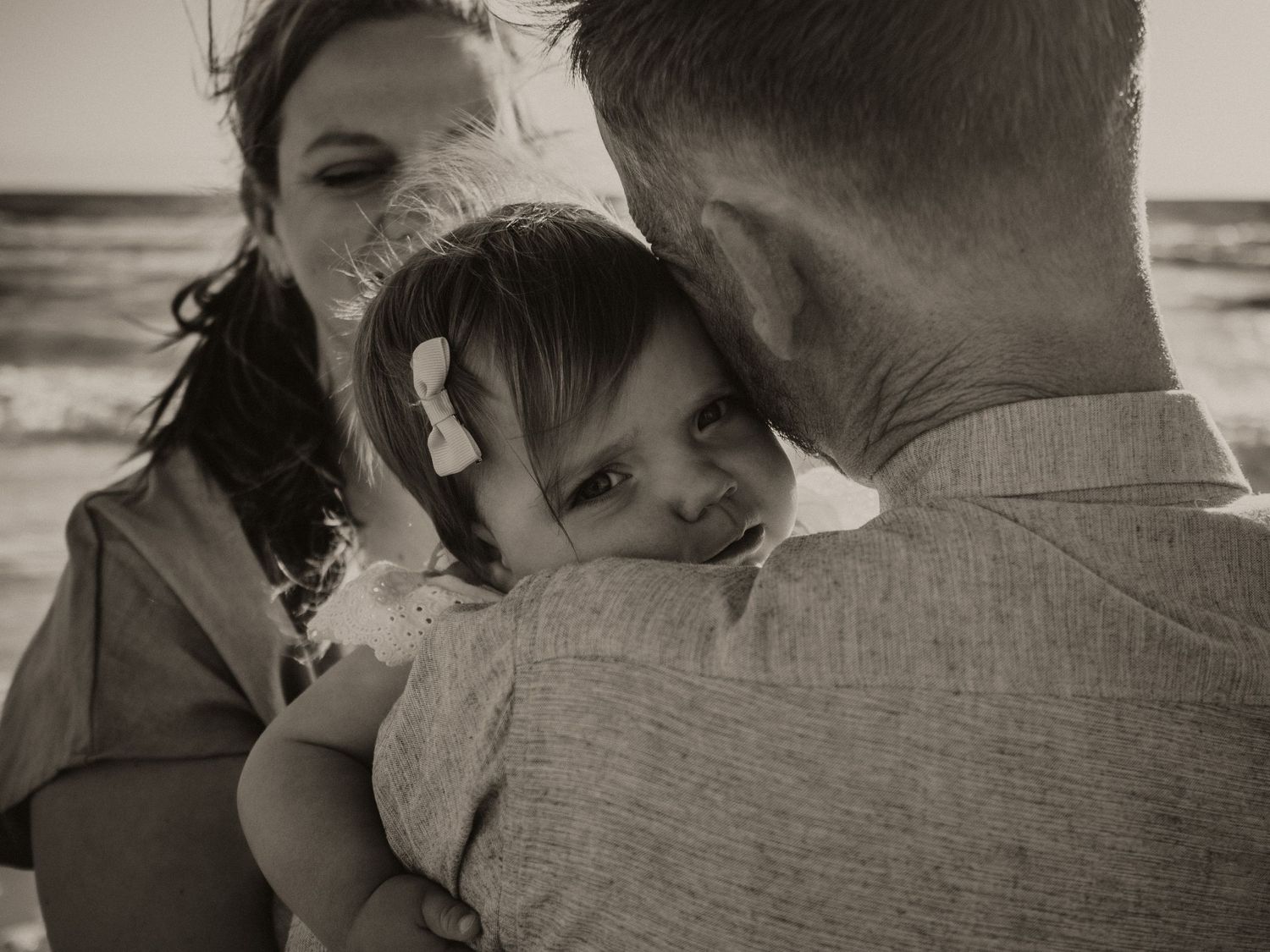 Baby girl being held over her fathers shoulder looking straight at the camera with the mother behind her