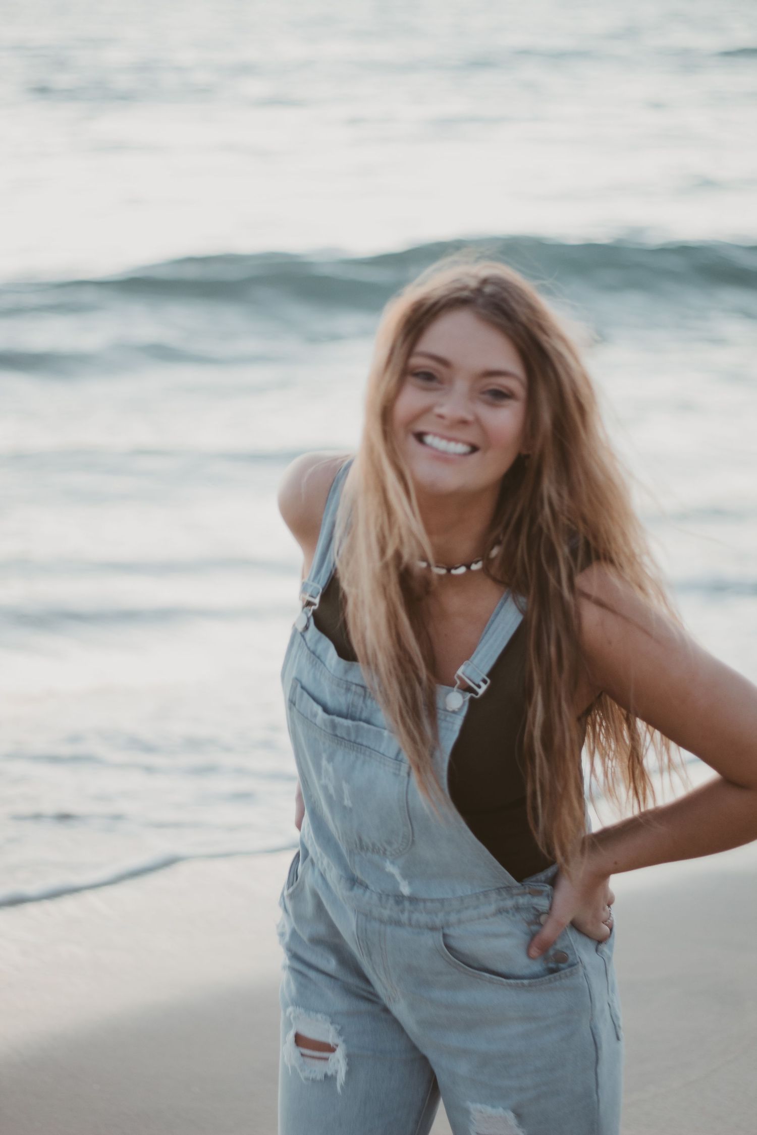 Young woman in denim overalls smiling at the beach with ocean waves in the background.