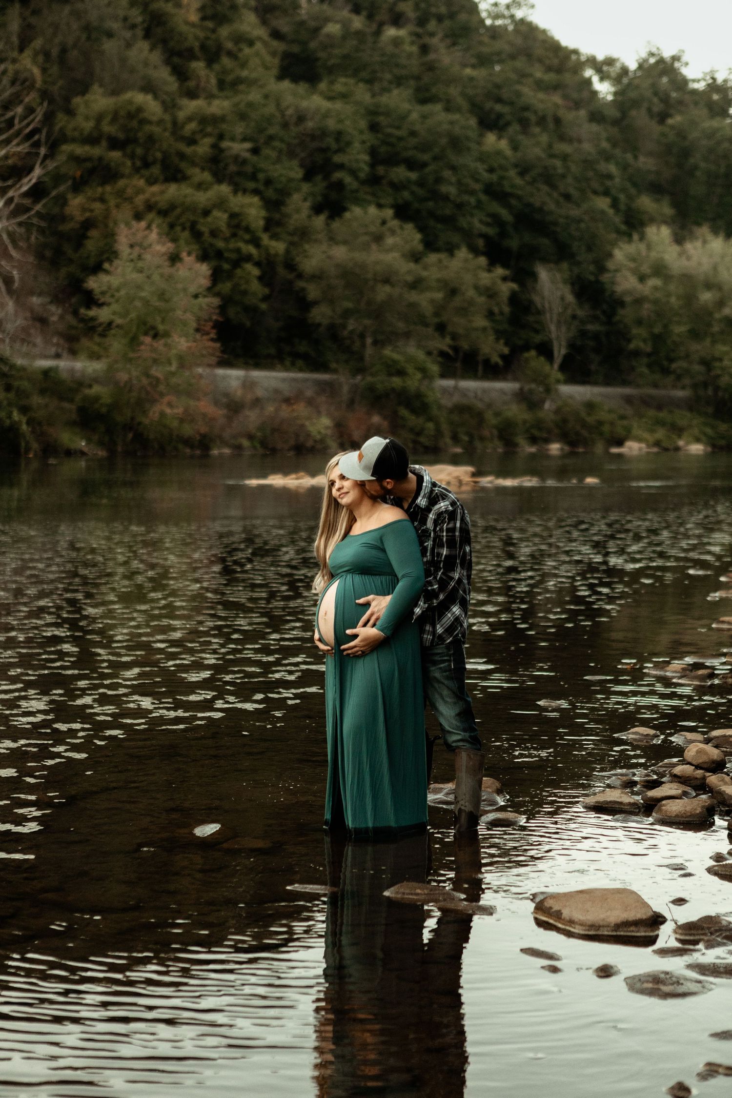 Maternity photo of couple standing in shallow river water during sunset.