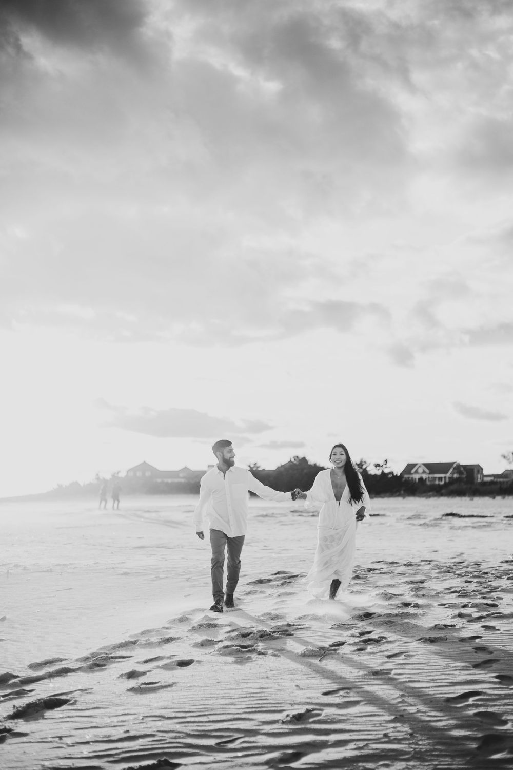 A couple walking on a misty beach at sunset holding hands in black and white photography.