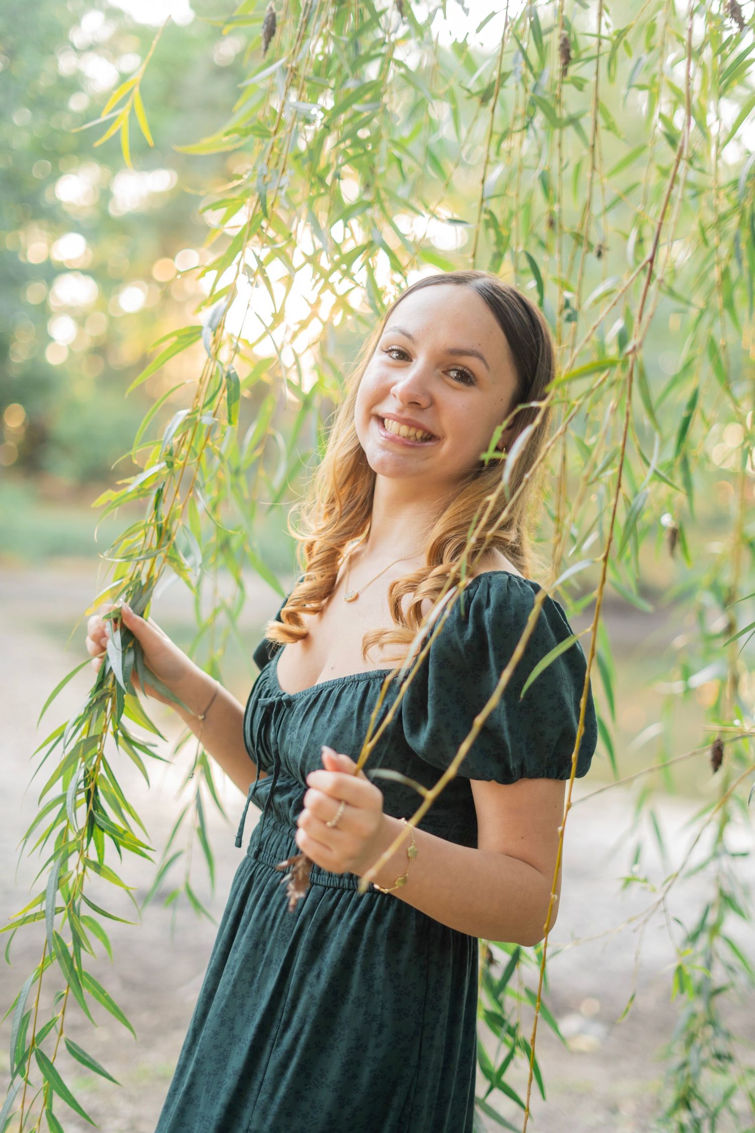Smiling young woman in green dress holding willow tree branches in golden sunlight.
