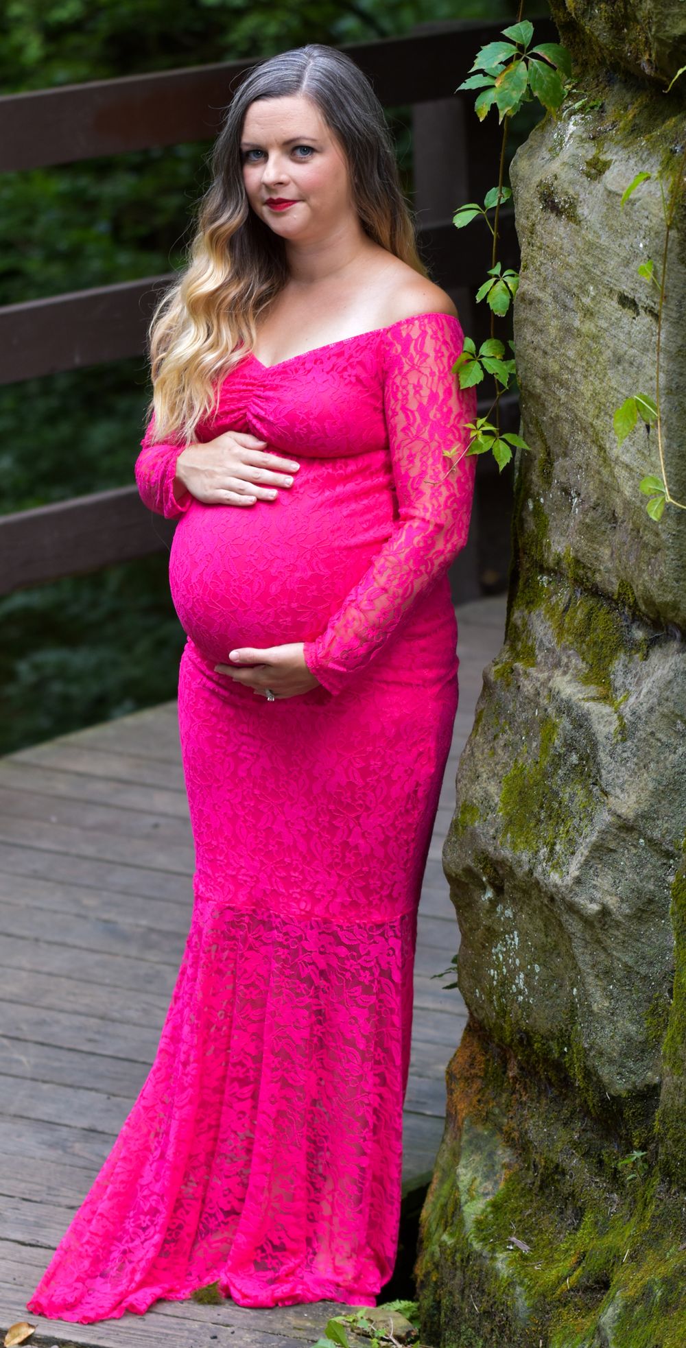 Expectant mother in a pink lace maternity gown poses outdoors by a mossy stone wall on a wooden deck.