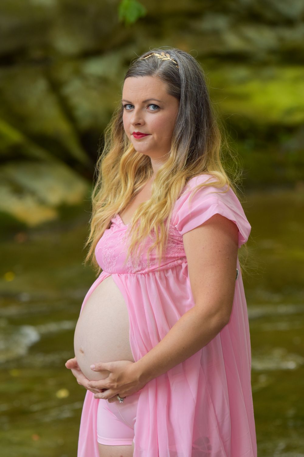 Maternity portrait in a flowing pink dress against a natural outdoor backdrop with green foliage.