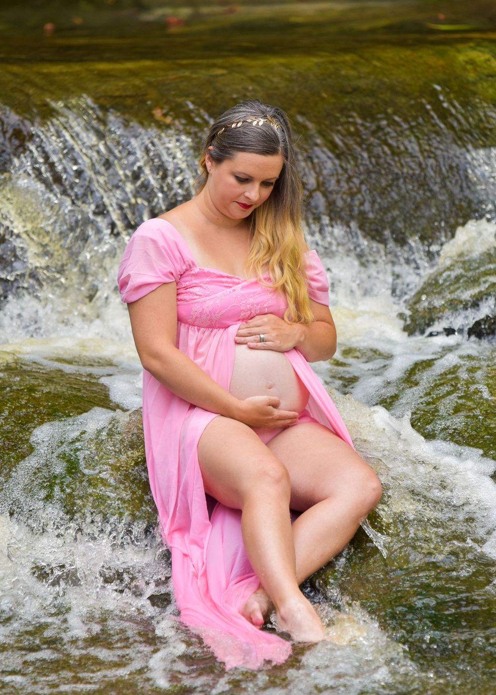 Maternity portrait in a pink dress sitting in flowing water with a waterfall background.