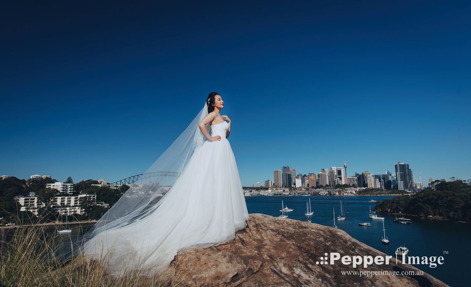 Bride in ball gown with long veil standing on rock overlooking Sydney Harbour skyline.
