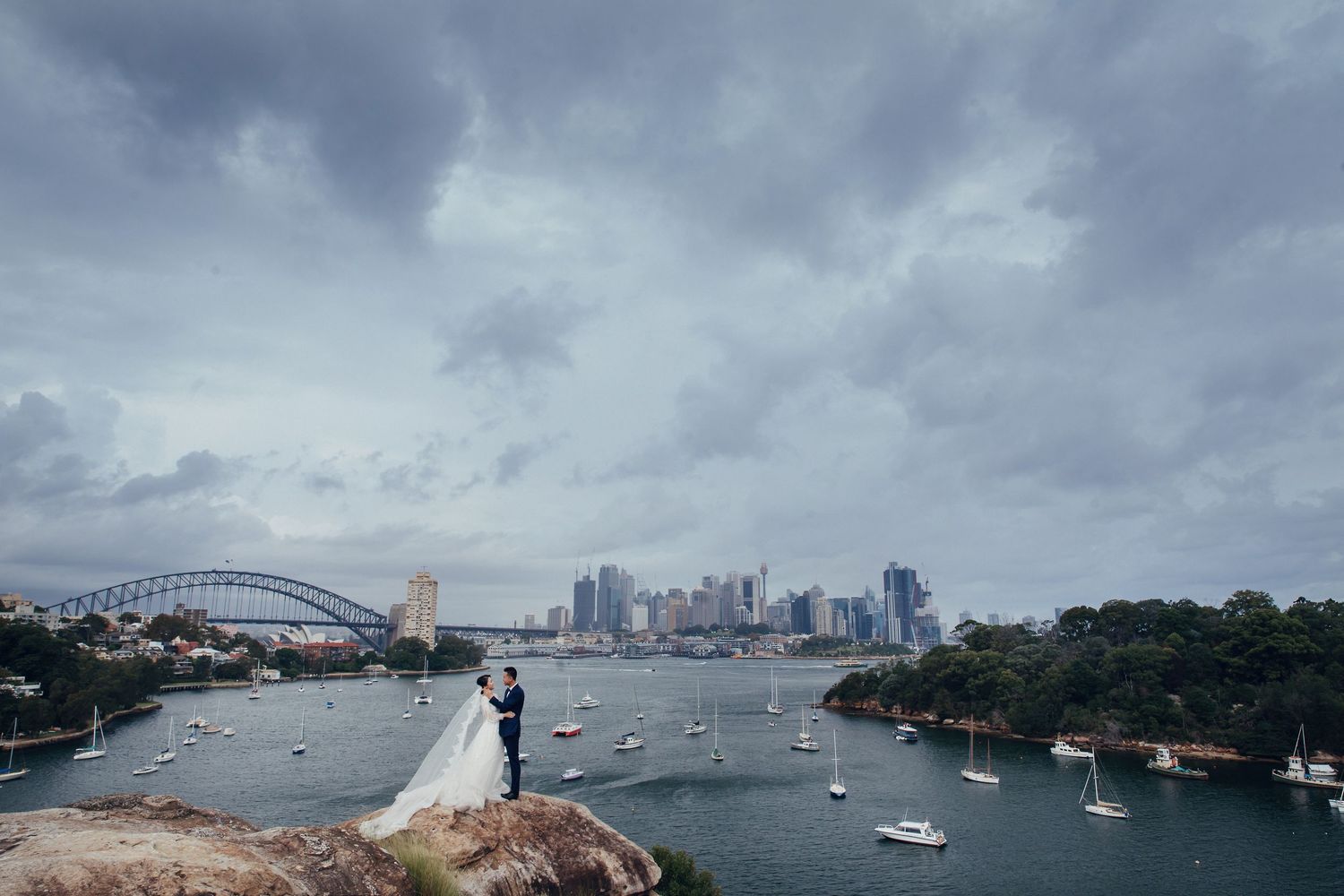 Wedding couple on rocky outcrop overlooking Sydney Harbour Bridge under dramatic cloudy sky.
