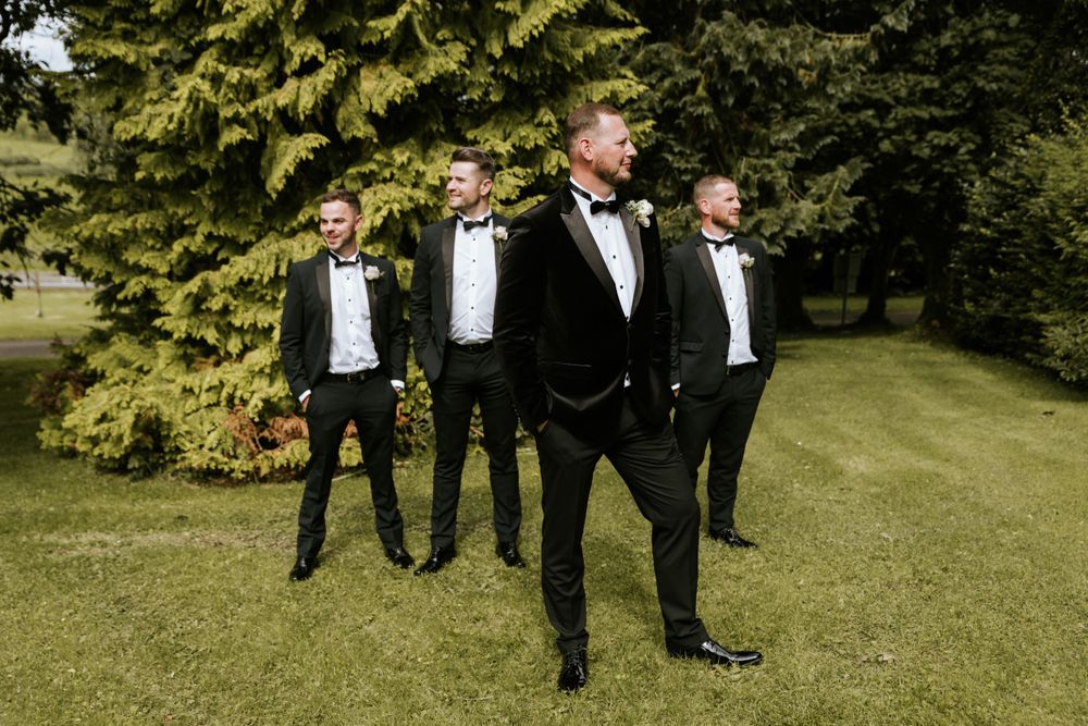 Wedding party in black tuxedos posing on green lawn with trees.