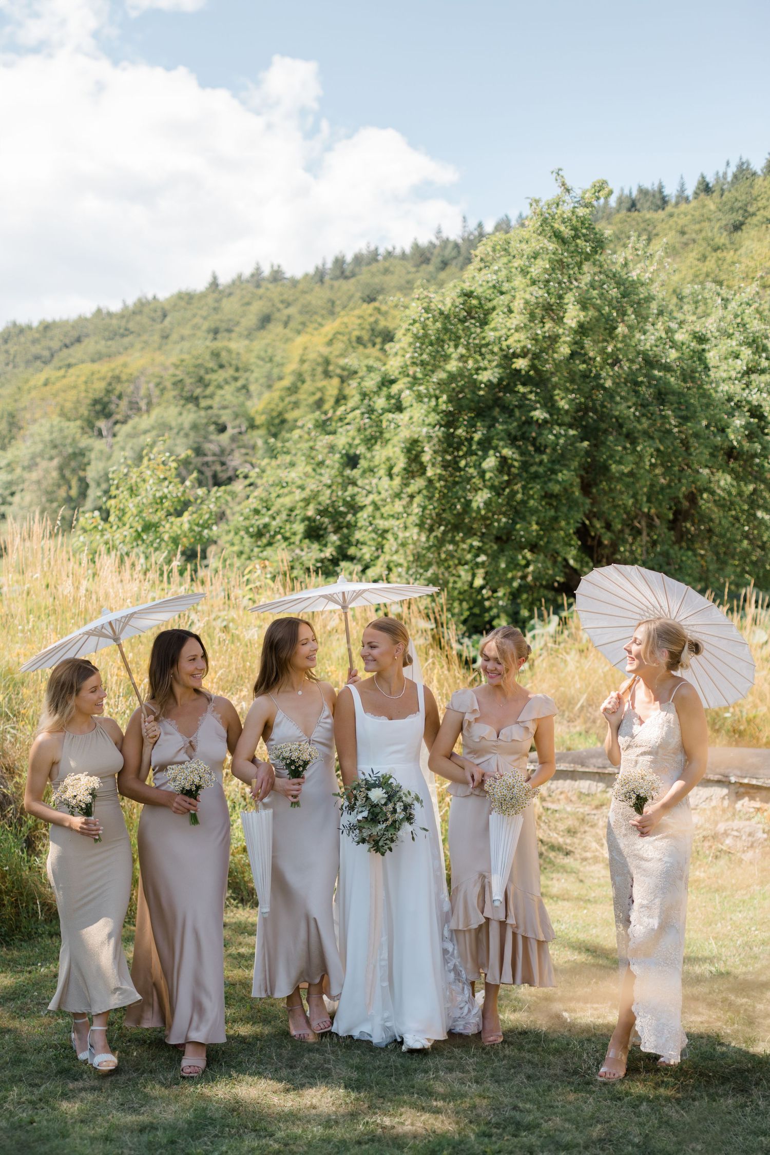 Bridal party in neutral dresses posing with parasols in outdoor garden setting.