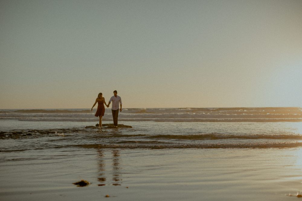 Silhouetted couple holding hands while walking along beach at sunset.