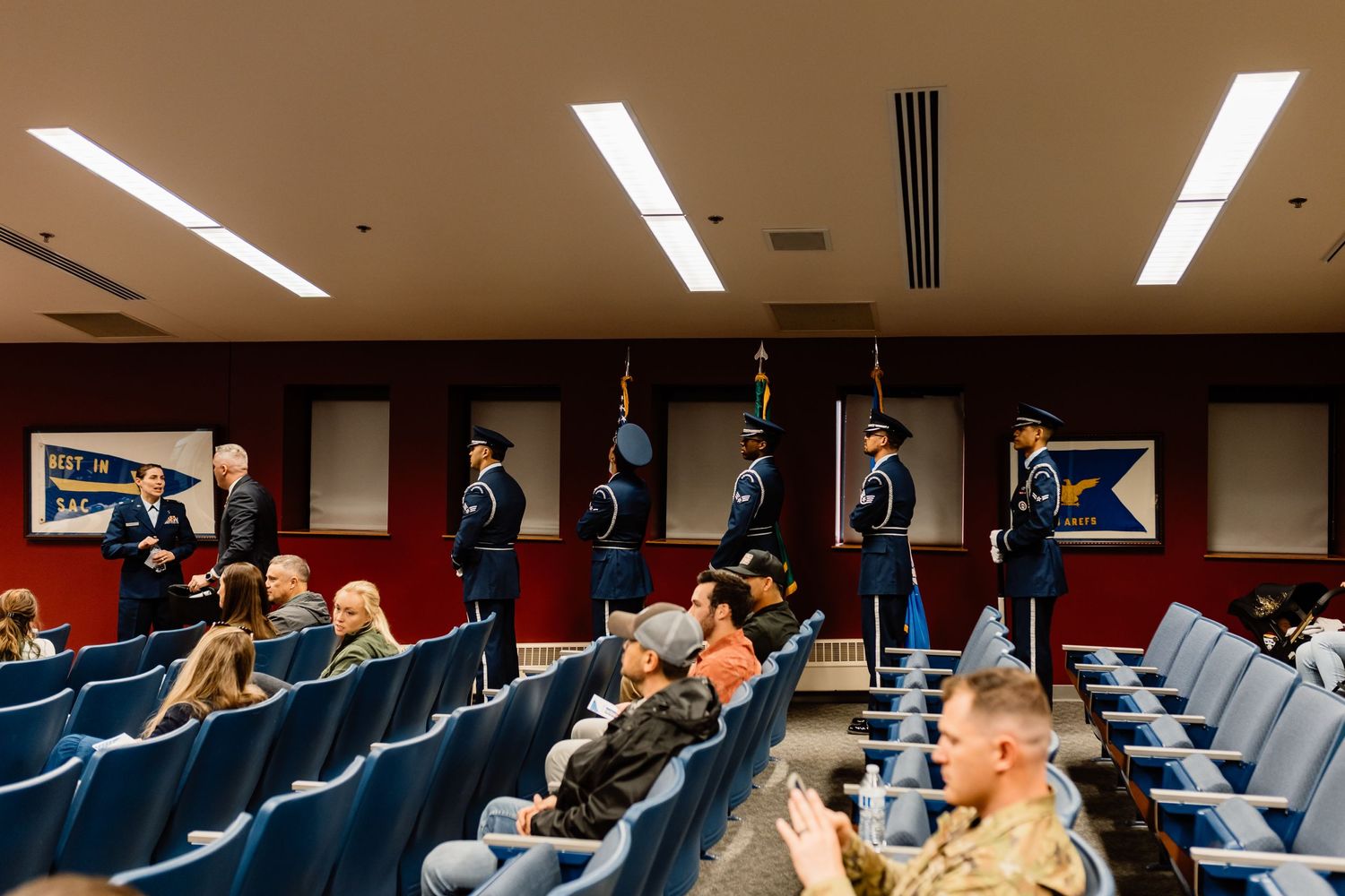 Military ceremony in auditorium with color guard and seated audience.