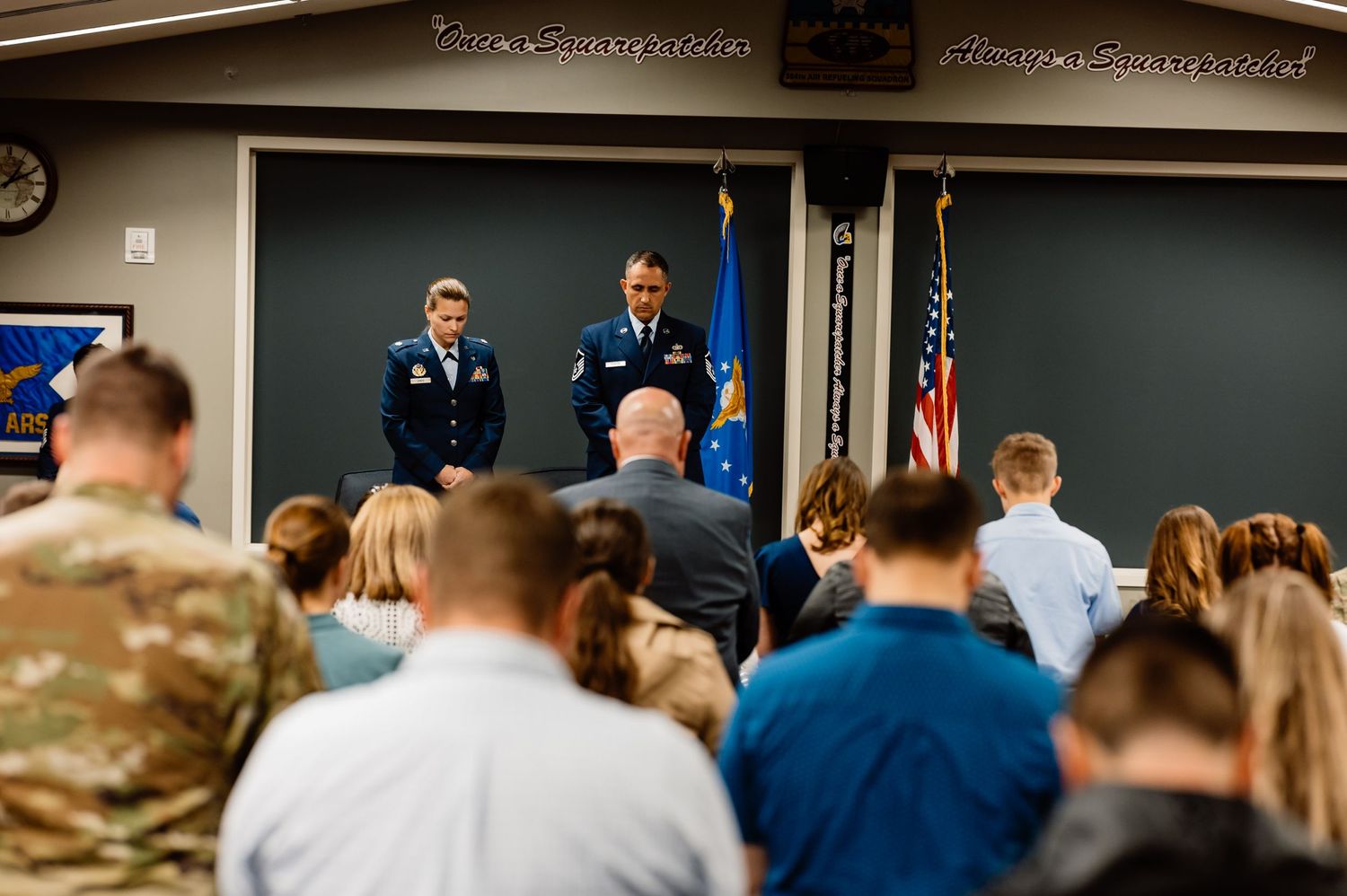 Military personnel and civilians gather in a classroom setting with American flags and officials at the front.