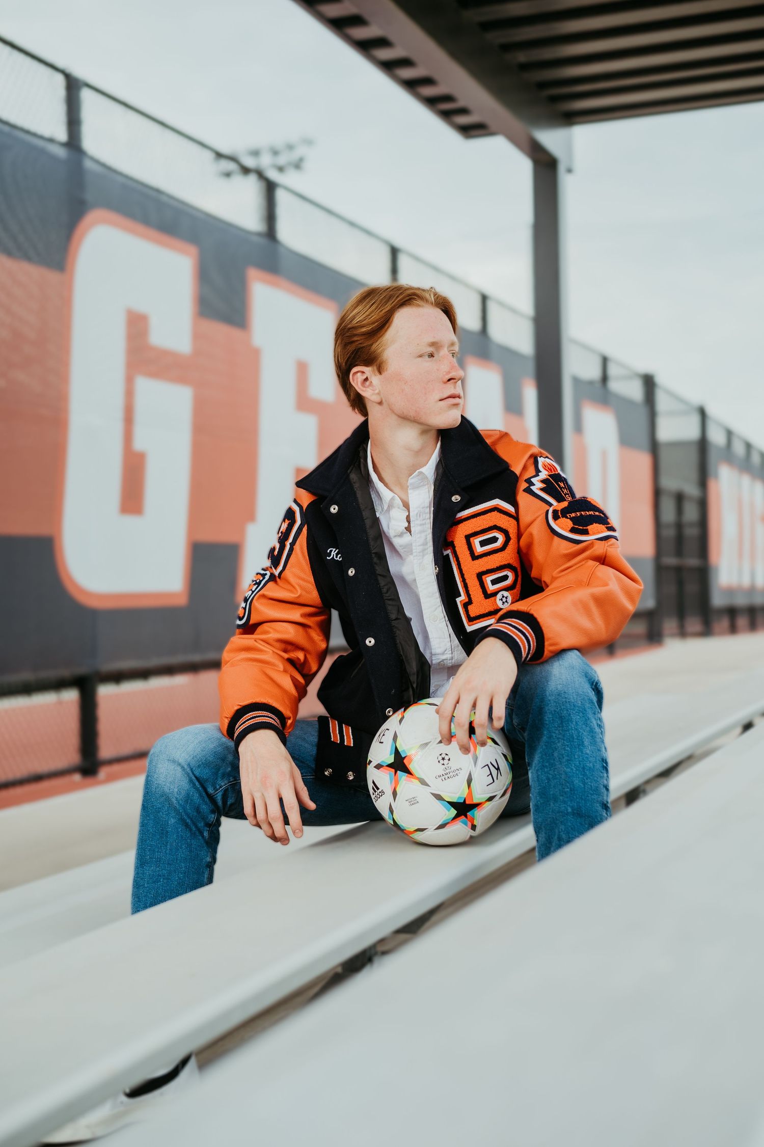 Person in varsity jacket sitting with soccer ball on bleachers.