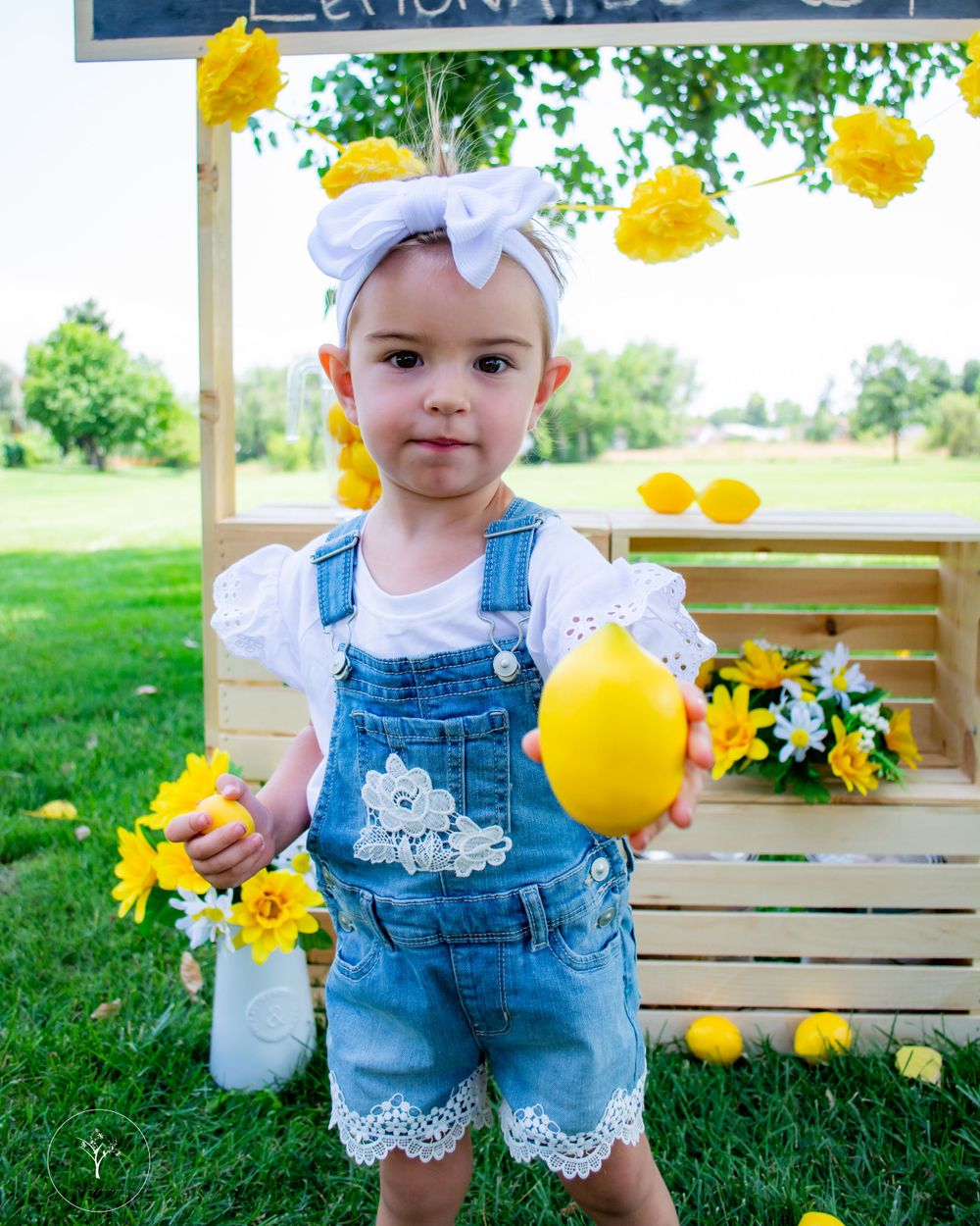 Child in denim overalls at sunny lemonade stand decorated with yellow flowers.