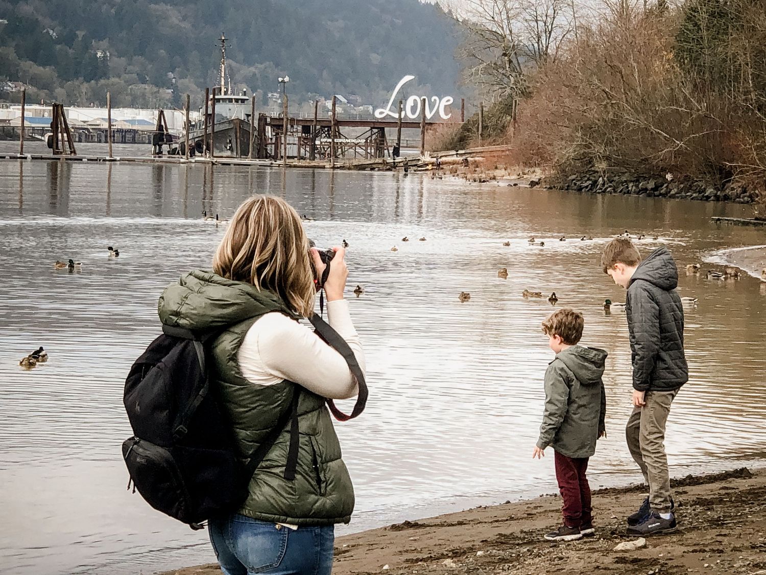 A photographer captures two children feeding ducks by a scenic riverside.