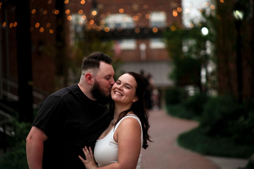 Couple sharing a tender kiss on a garden path with warm string lights glowing softly in background.