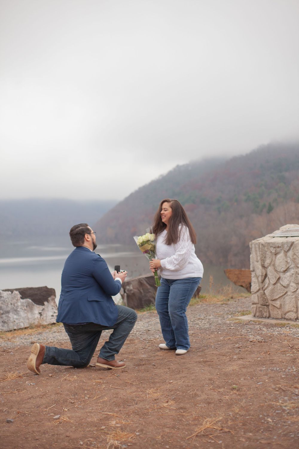 Man on one knee proposing to a woman with flowers on a misty overlook above a lake and mountains.