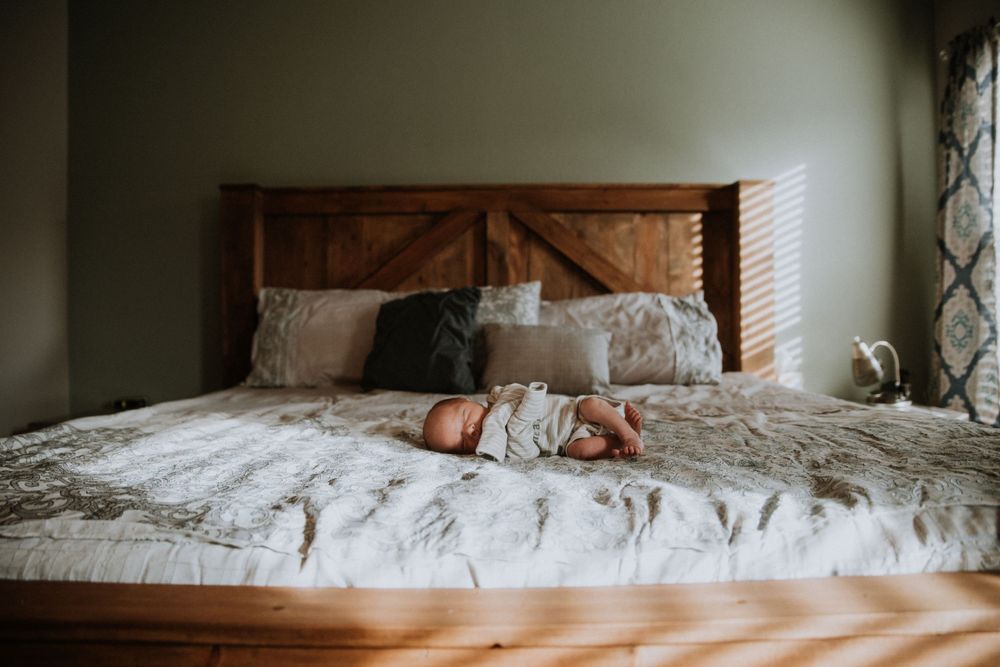 Newborn baby peacefully resting in the center of a bed, bathed in soft natural light streaming through the window, creating a soft moment.