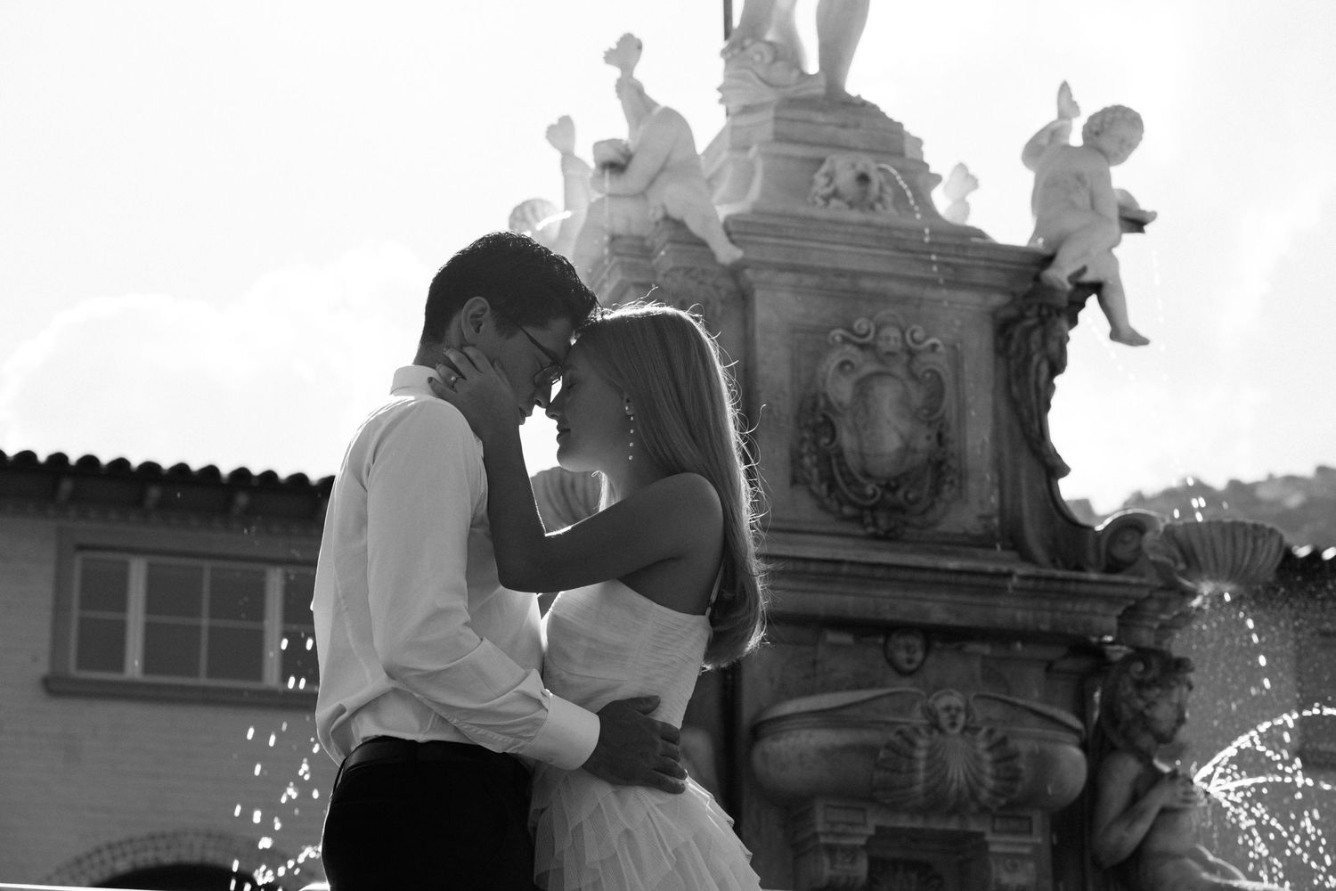 Black and white photo of couple embracing near ornate fountain with twinkling lights.