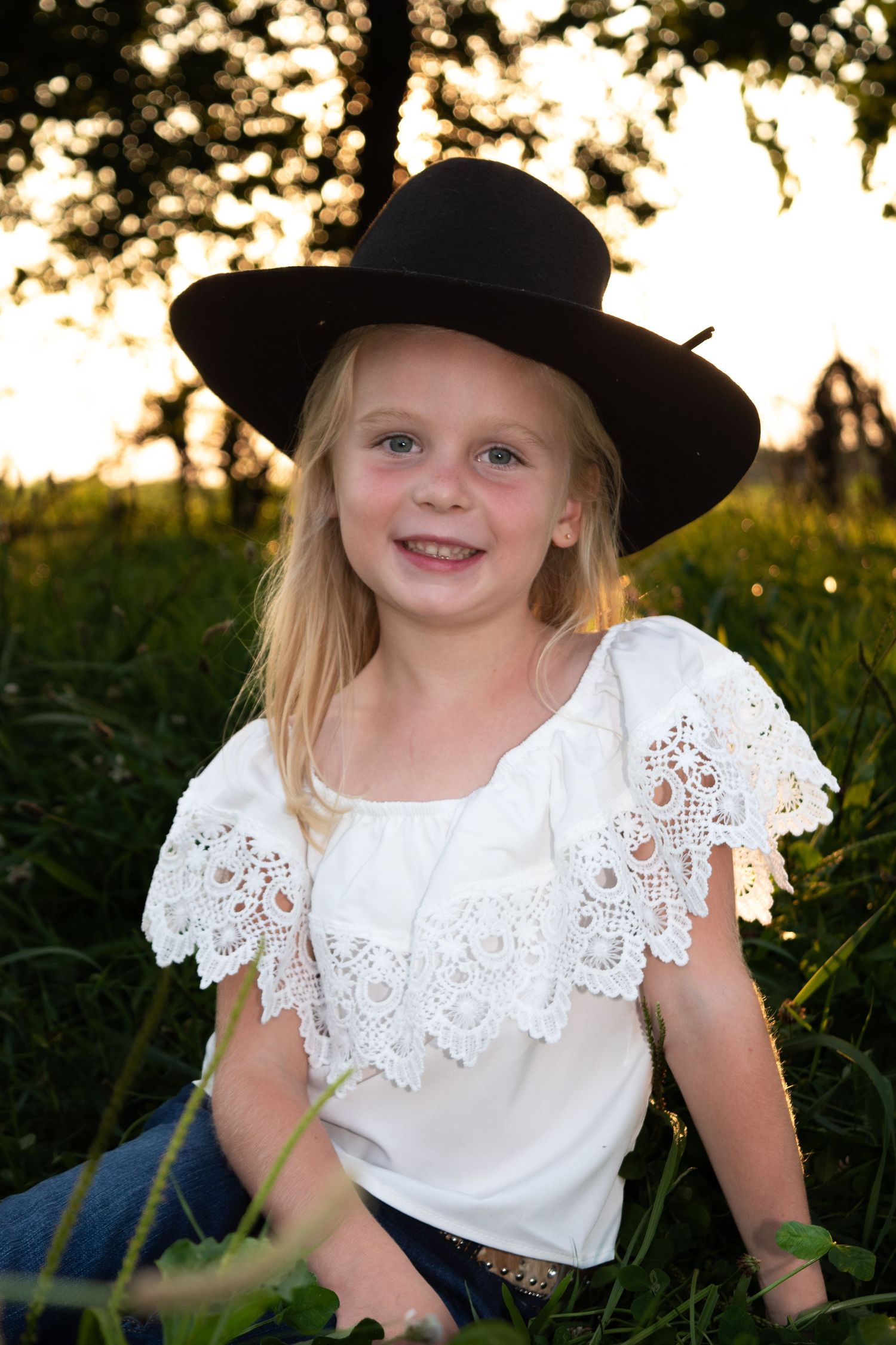 A young person wearing a black hat and white lace top poses outdoors at golden hour.