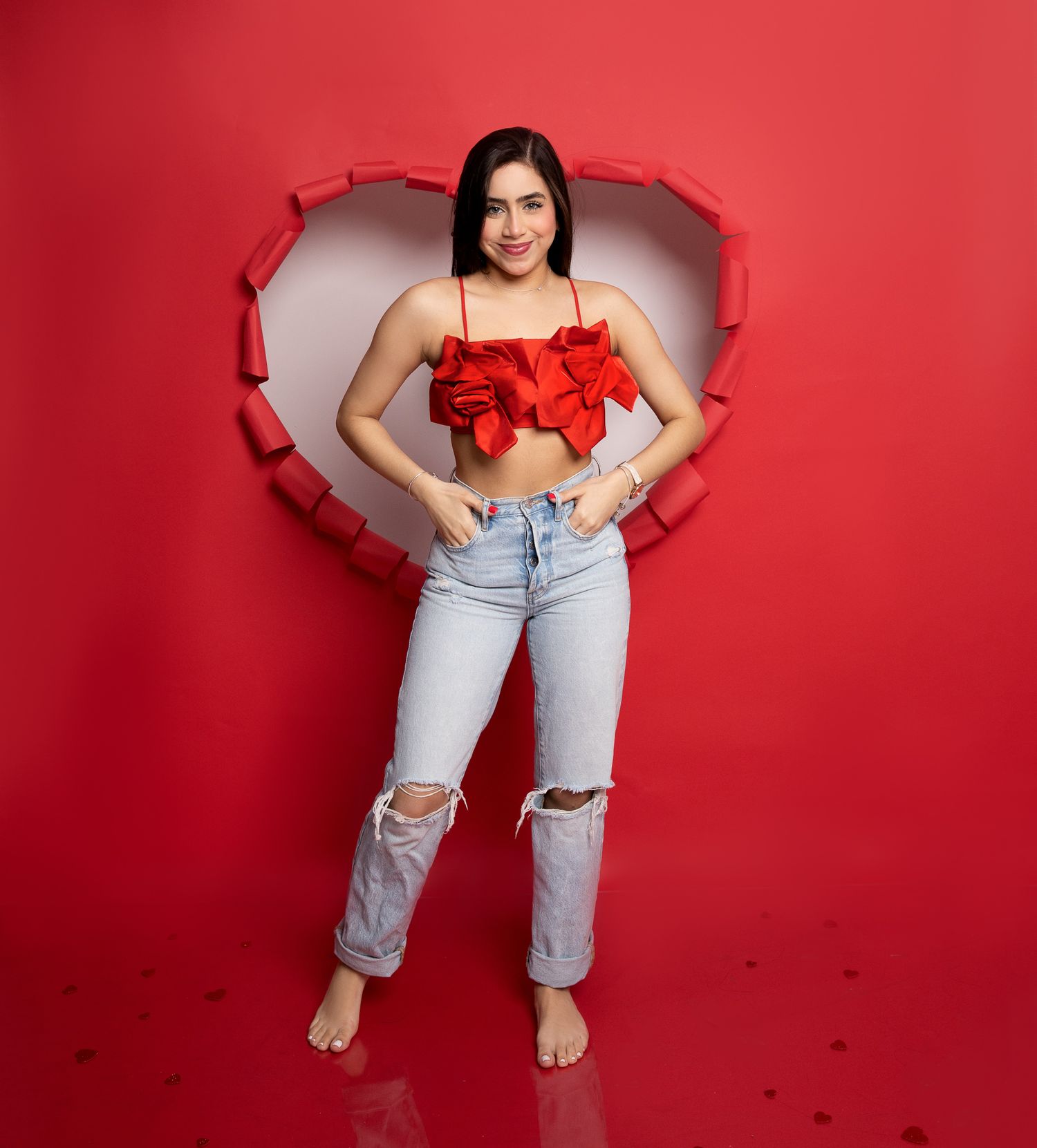 A person posing in ripped jeans and red top against a heart-shaped cutout in red background.