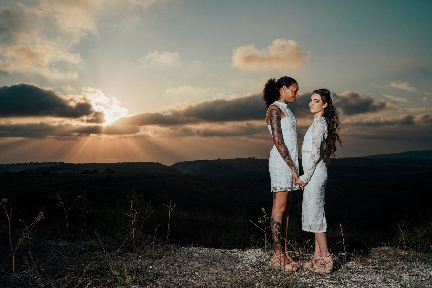 A couple in white outfits stands together against a stunning mountain sunset.