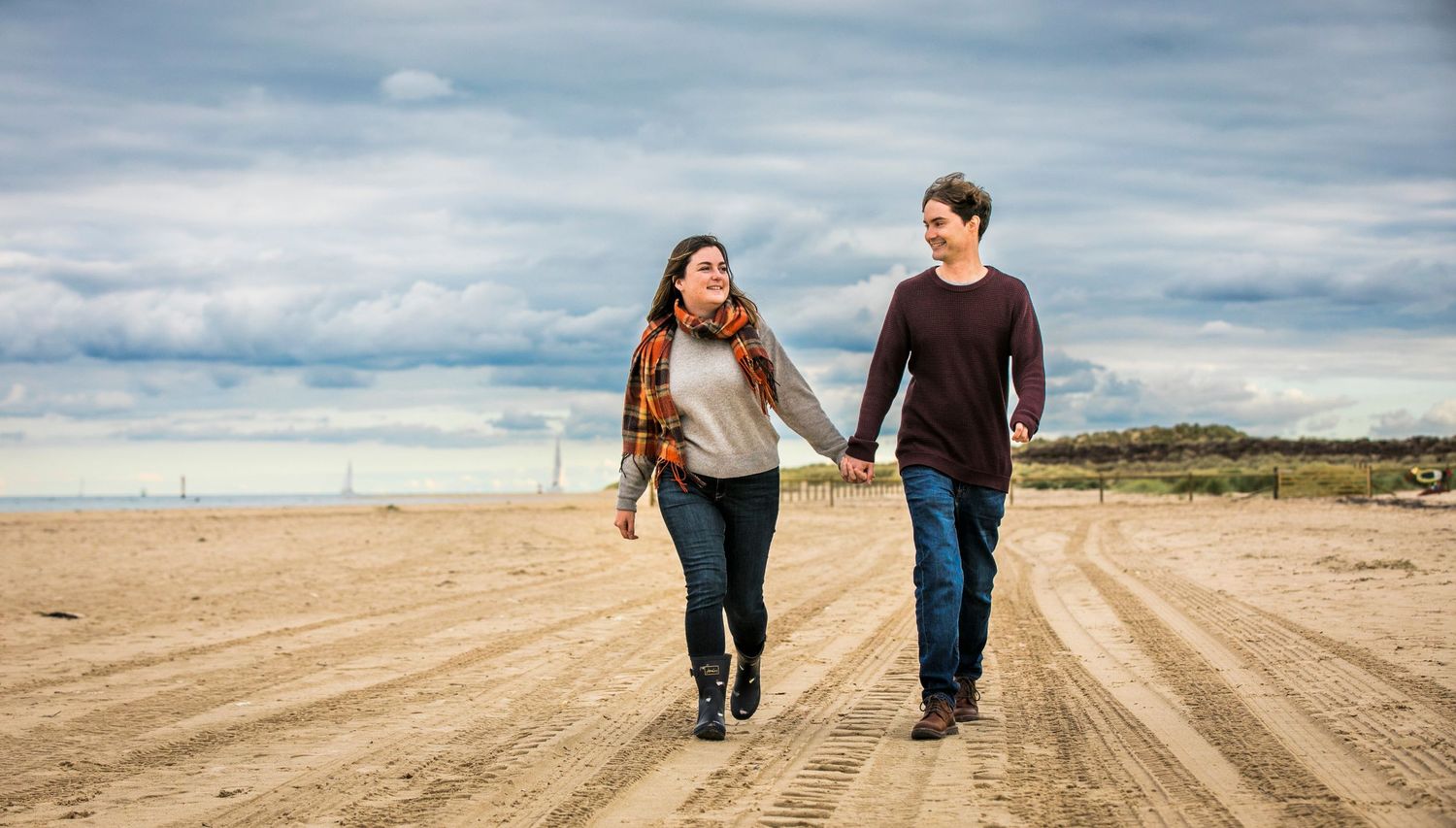 A couple walking hand in hand on a wide sandy beach path under cloudy skies.