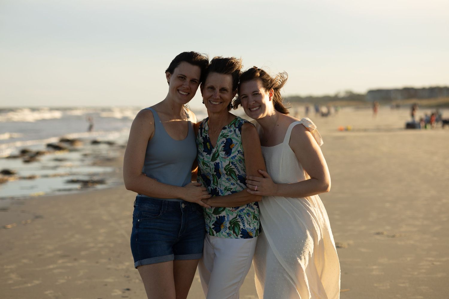 Three figures embrace together on a sunlit beach at sunset.